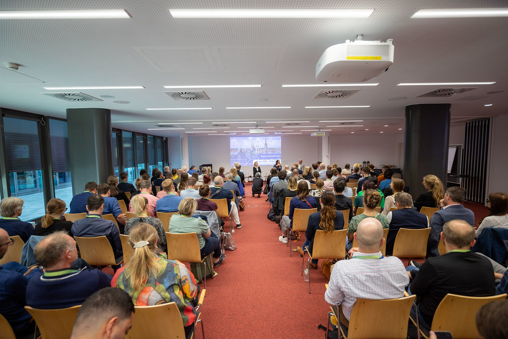 Menschen sitzen in einem Raum und schauen nach vorn auf einen Präsentation