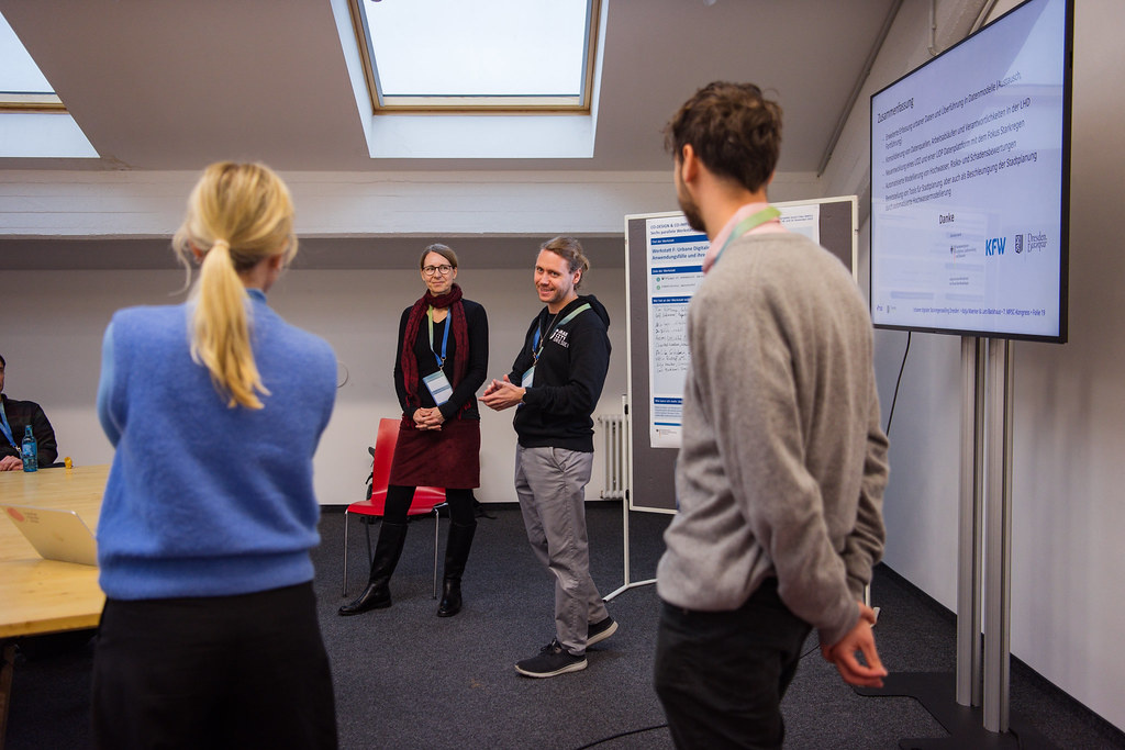 In the foreground, two moderators seen from behind, with the two speakers in front of them.